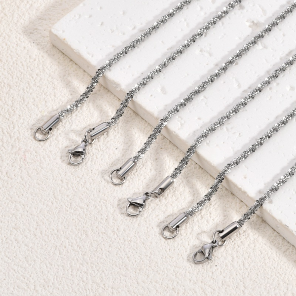 Finished stainless steel sparkle chain necklaces with lobster clasps laid neatly on a white textured stone surface for B2B supplier web site.