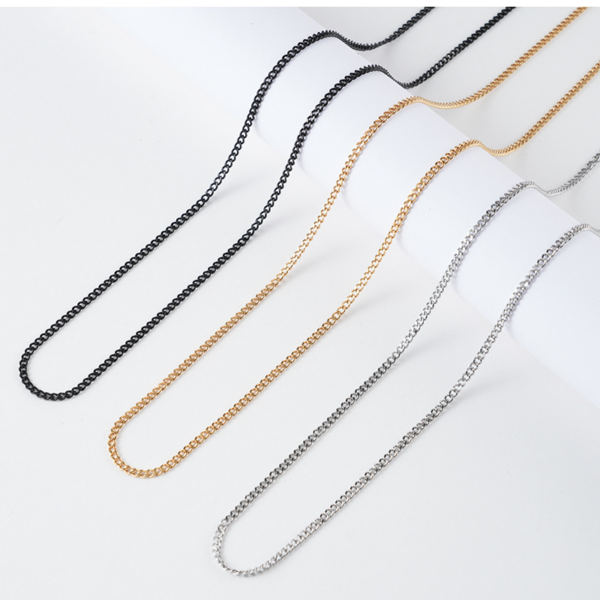 Finished stainless steel curb chain necklaces in matte black, 18K gold, and polished silver draped elegantly on a white cylindrical display step.