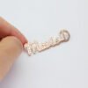 满钻字母胸针 (1) A close-up view of a personalized rose gold stainless steel cursive letter name brooch pin held by fingers against a neutral backdrop.