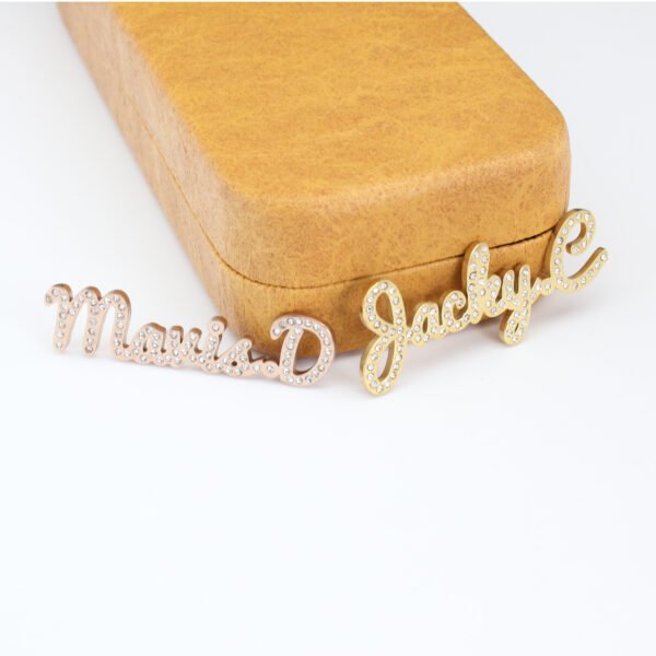 满钻字母胸针 (5) Parallel display of wholesale custom rhinestone name brooches in rose gold and yellow gold finishes leaning against a jewelry gift box.