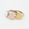 圆牌戒指_0002_图层 7 Three plain, round rings—in silver, gold, and rose gold—are arranged in an overlapping pattern on a solid-colored background, showcasing their exquisite craftsmanship.