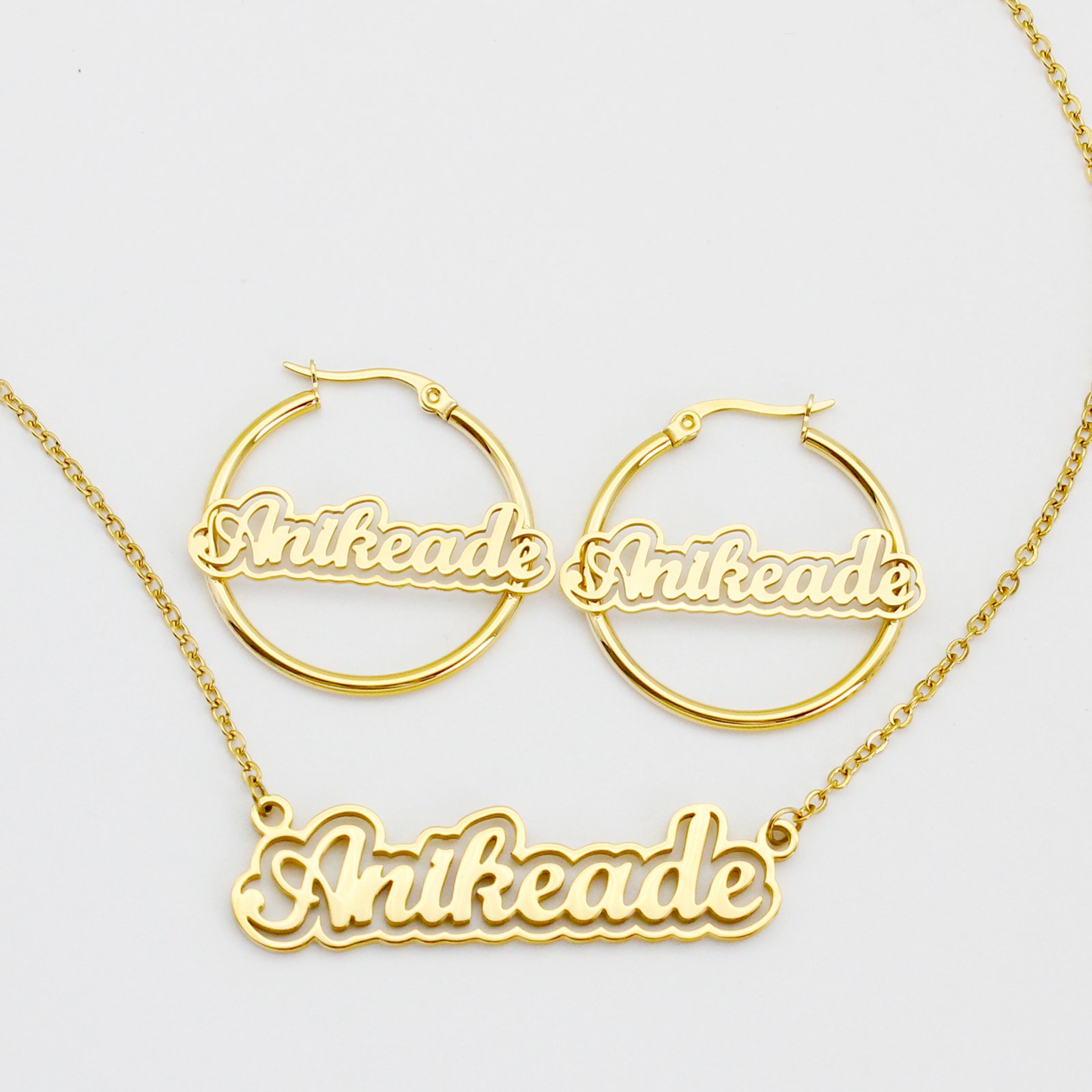 A blank finished 18K gold plated stainless steel personalized 2-layer cursive name necklace and hoop earrings set spelling "Anikeade" on a white background.