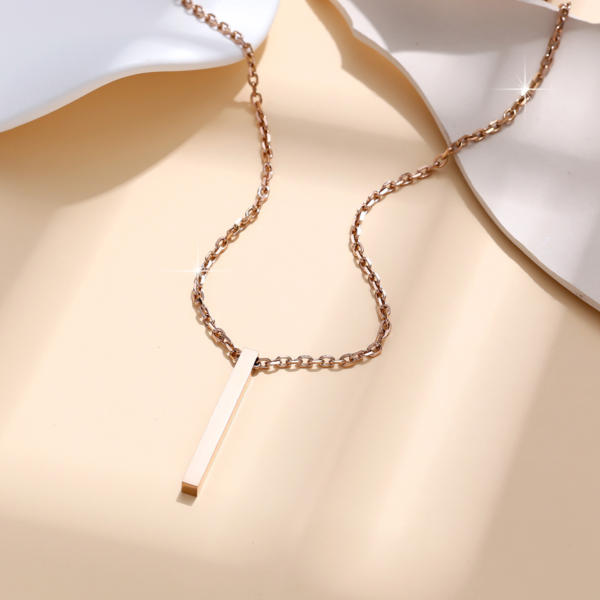A blank finished rose gold plated stainless steel rectangle pillar pendant necklace draped fluidly on a warm beige textured backdrop for B2B catalog.