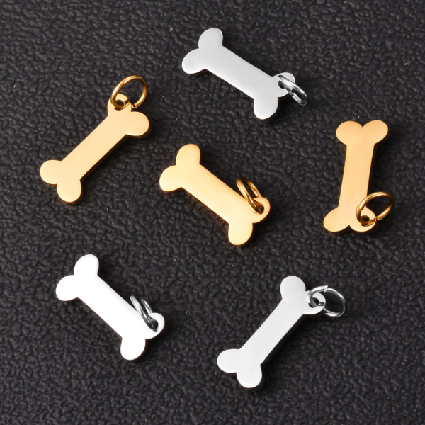 An assortment of small silver and gold stainless steel bone charms with mini jump rings displayed on a textured dark grey leather-like background.